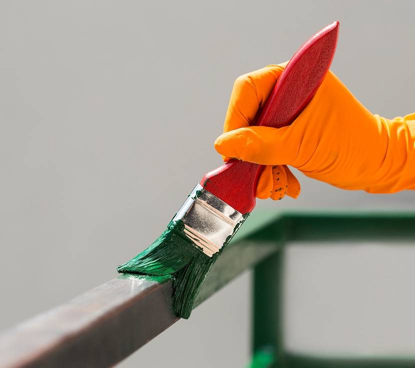 Eine Person, die einen orangenen Handschuh trägt, streicht mit einem roten Pinsel ein Geländer grün an.