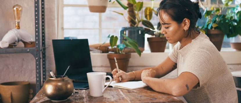 Frau sitzt am Schreibtisch neben Laptop und skizziert etwas auf einem Notizblock.