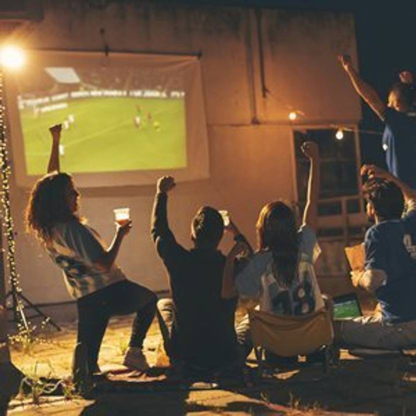 Menschen feiern vor Beamer Leinwand, auf der Fußball läuft