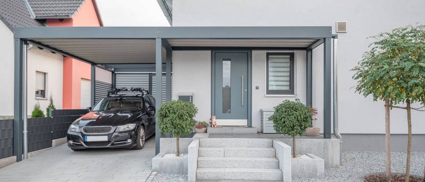 Ein Carport inklusive Überdachung vor der Haustür eines weißen Hauses. Unter dem Carport wurde ein schwarzes Auto geparkt.