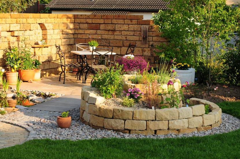 Ein Garten mit einem hochgebauten Beet, das von einer Steinmauer umgeben ist. Im Hintergrund wurde eine Sitzecke eingerichtet.