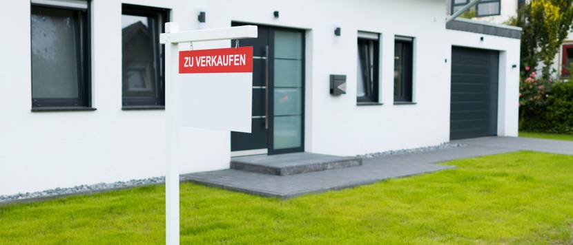 Vor einem weißen Haus mit gepflegten Vorgarten steht ein Schild mit der Aufschrift