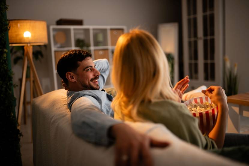 Ein Paar sitzt auf dem Sofa. Im Hintergrund steht eine Stehlampe, die warmes Licht ausstrahlt.