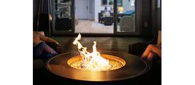 Auf einer Terrasse vor einem Haus brennt in einer Feuerschale ein Feuer. Zu beiden Seiten befinden sich Beine von sitzenden Personen.