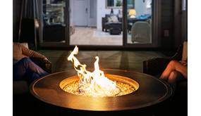 Auf einer Terrasse vor einem Haus brennt in einer Feuerschale ein Feuer. Zu beiden Seiten befinden sich Beine von sitzenden Personen.
