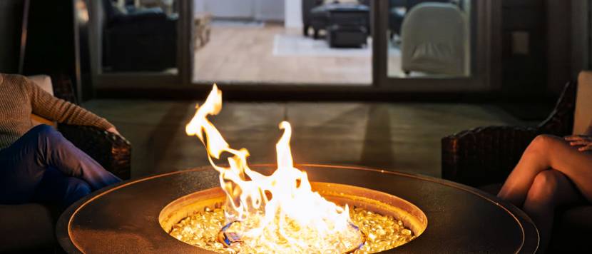 Auf einer Terrasse vor einem Haus brennt in einer Feuerschale ein Feuer. Zu beiden Seiten befinden sich Beine von sitzenden Personen.