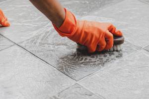 Hand mit orangenem Handschuh schrubbt mit einer Bürste den Fliesenboden.