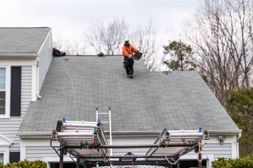 Eine Person in Arbeitskleidung deckt ein Haus neu ein, im Vordergrund steht ein Baufahrzeug