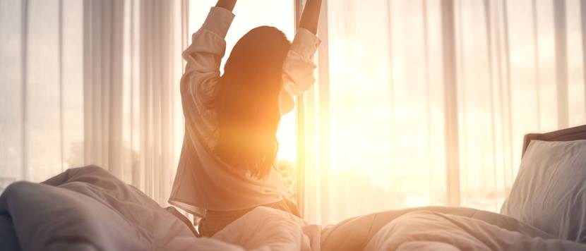 Frau streckt sich im Bett. Im Hintergrund geht die Sonne auf.