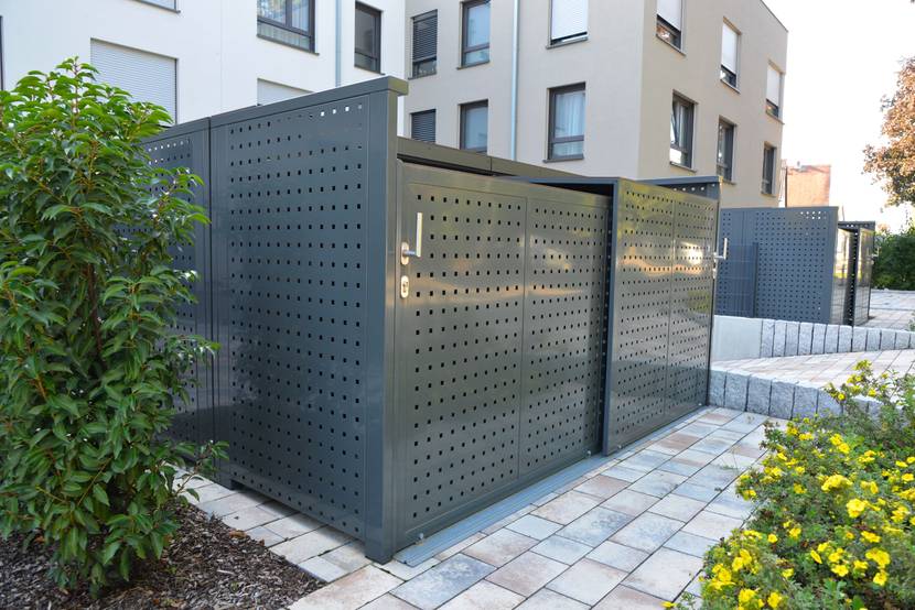 Mülltonnenbox aus Metall steht vor einem Mehrfamilienhaus.