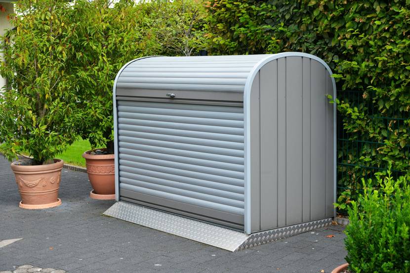 Eine graue Mülltonnenbox steht vor einer Hecke.