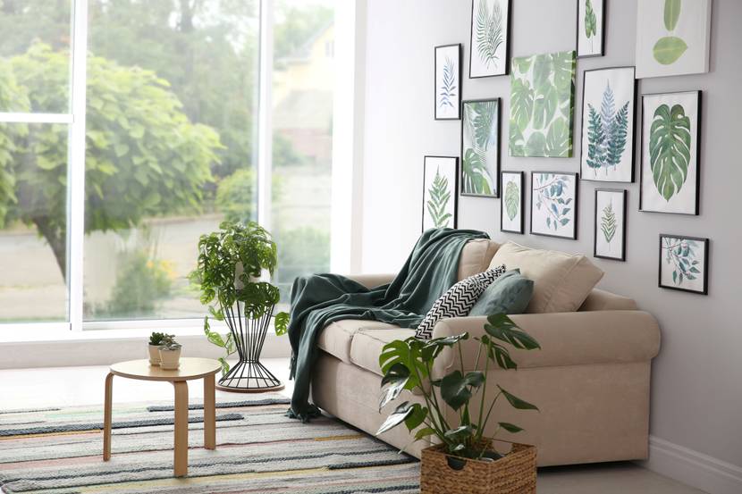 In einem modernen Wohnzimmer mit großer Fensterfront steht ein helles Sofa, hinter dem eine Bildergalerie mit Pflanzenmotiven hängt.
