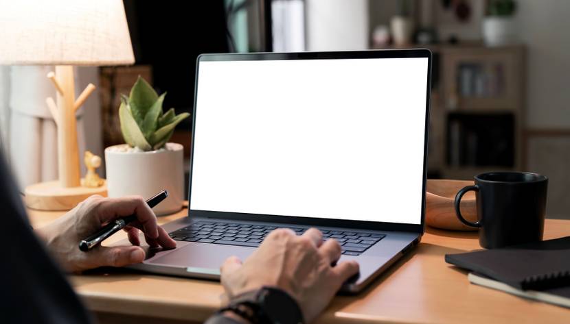Ein Mann arbeitet an einem Laptop. Auf seinem Schreibtisch stehen eine Schreibtischlampe, eine schwarze Tasse und eine Pflanze.
