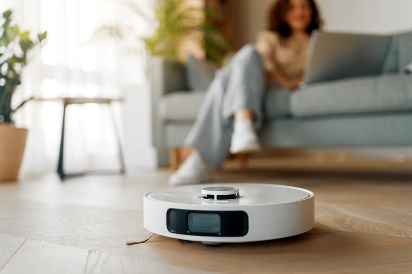Ein Staubsaugerroboter saugt ein Wohnzimmer, während im Hintergrund eine Frau auf dem Sofa sitzt und am Laptop arbeitet.
