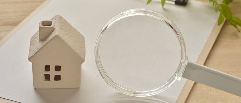 Auf einem Holztisch liegt ein Klemmbrett mit leerem Papier. Auf dem Klemmbrett wurde ein Modellhaus und eine Lupe platziert.