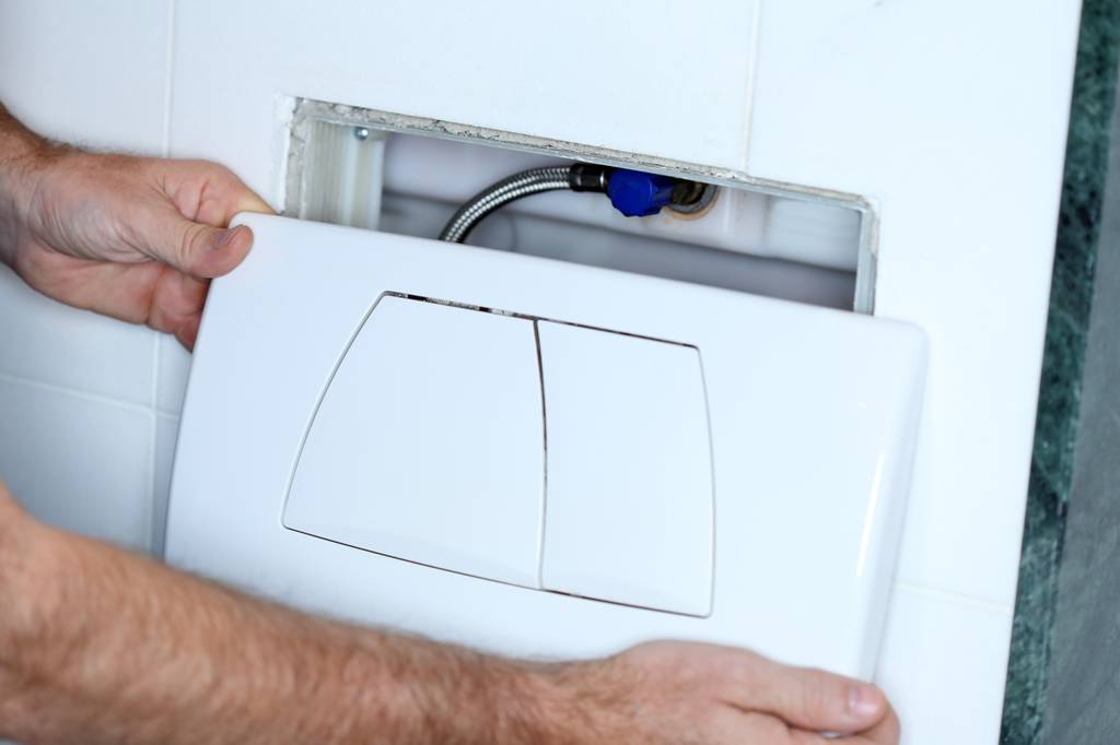 Hände montieren eine Betätigungsplatte vor einem Unterputz-Spülkasten, der durch die geöffnete Wandöffnung sichtbar ist.