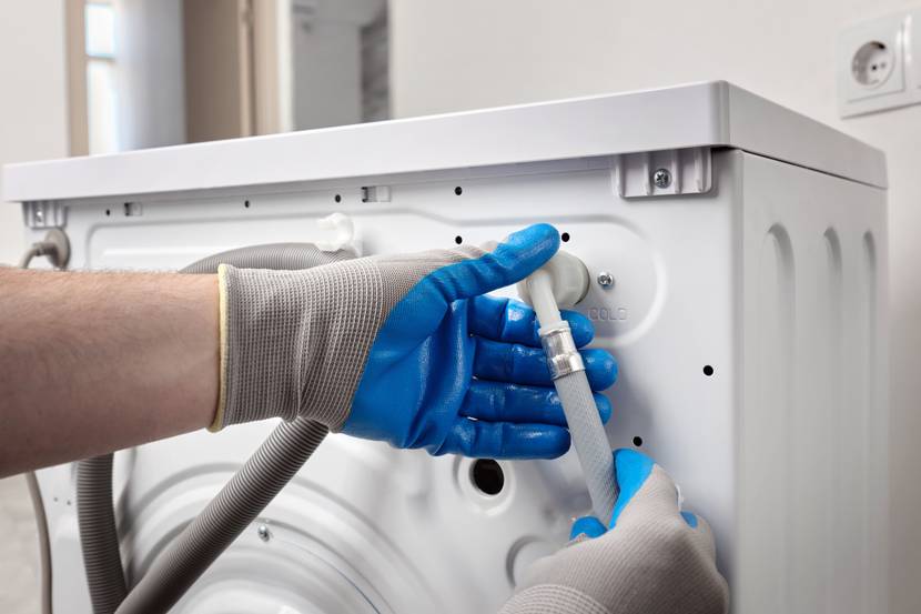 Hände mit Arbeitshandschuhen prüfen einen Schlauchanschluss an der Rückseite einer Waschmaschine.