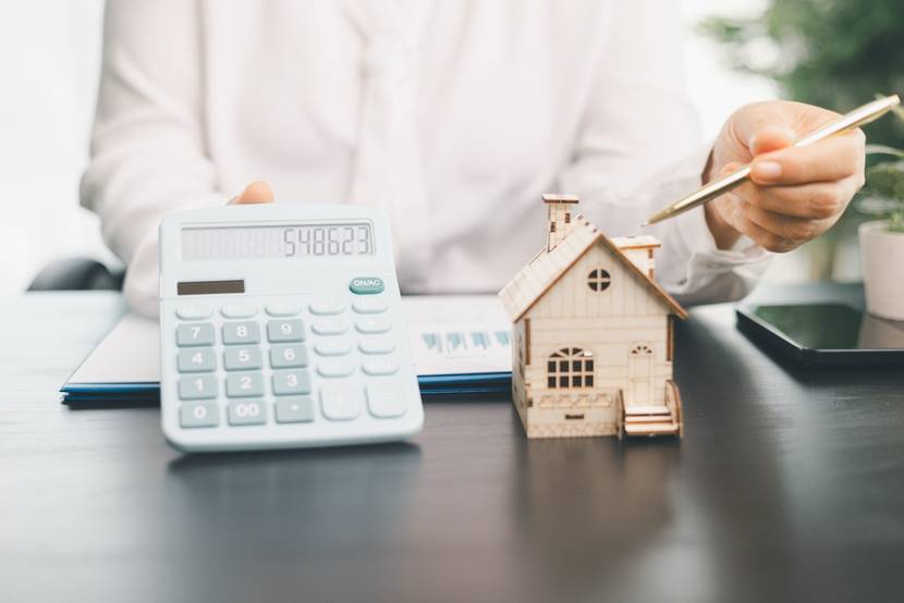 Ein Schreibtisch mit einem großen Taschenrechner und einem Haus-Modell, auf das die Person hinter dem Schreibtisch deutet.