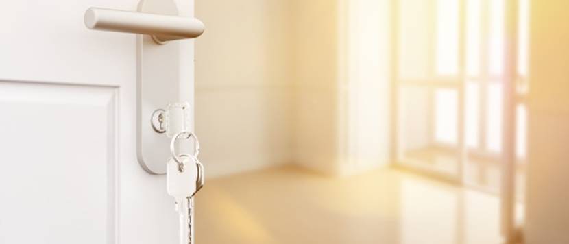 In einer offenstehenden Tür steckt ein Wohnungsschlüssel. Im Hintergrund befindet sich eine leerstehende Wohnung.
